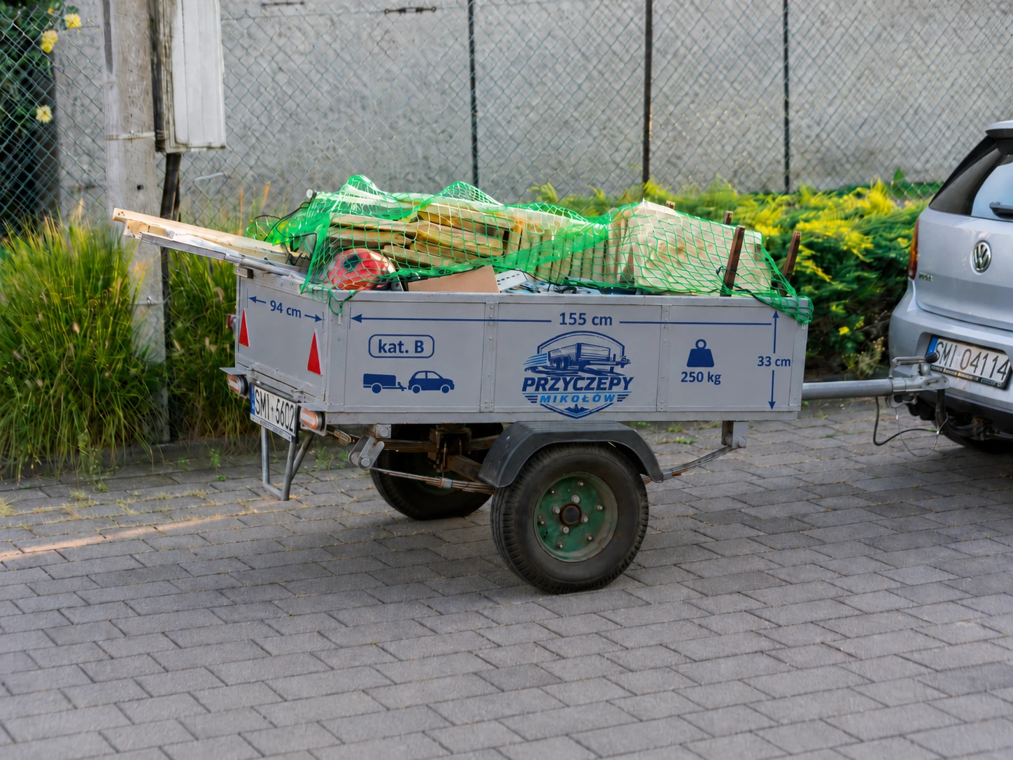 Przyczepa S-250 z ładunkiem pod siatką, oznaczeniami wymiarów 155 × 94 × 33 cm i ładownością 250 kg